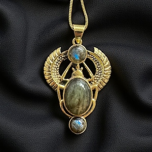 Peut inclure: Un collier pendentif doré avec un design de scarabée égyptien. Le pendentif présente une pierre centrale ovale et deux petites pierres rondes, avec des détails d'ailes. Le collier est présenté sur un fond noir.