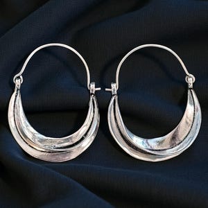 Peut inclure: Une paire de boucles d'oreilles créoles argentées. Chaque boucle d'oreille présente un design en forme de croissant avec des détails superposés. Les boucles d'oreilles ont un style simple et élégant et sont suspendues à un fil incurvé. Le fond est un tissu sombre et texturé.