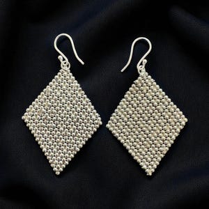 Peut inclure: Une paire de boucles d'oreilles argentées en forme de losange. Chaque boucle d'oreille est composée de petites perles rondes, créant une surface texturée. Les boucles d'oreilles ont une fermeture à crochet simple, sur fond noir.