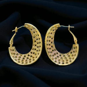 Peut inclure: Une paire de boucles d'oreilles créoles dorées. Les boucles d'oreilles présentent un motif ajouré texturé avec un motif répétitif de petits carrés. Elles sont posées sur un fond bleu foncé.