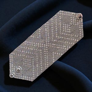 Può includere: Un braccialetto argentato con un design a perline. Il braccialetto rettangolare ha angoli arrotondati e una chiusura a bottone. Lo sfondo è un tessuto blu scuro.