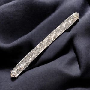 May include: A silver-toned mesh bracelet featuring a fine, textured chain-link pattern. The bracelet is finished with two circular, embossed metal studs at one end and a smooth, rounded metal clasp at the opposite end, creating a sleek design.