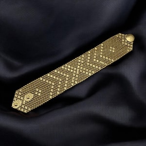 May include: A gold-colored cuff bracelet with a woven pattern and two button closures. The bracelet is rectangular with rounded edges and is displayed on a dark blue satin surface. The bracelet is made of small, interconnected metal pieces.