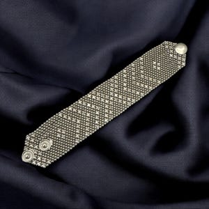 Op de afbeelding: Zilverkleurige armband met een geweven, getextureerd ontwerp. De armband heeft een reeks kleine, onderling verbonden vierkanten, waardoor een gedetailleerd patroon ontstaat. Hij sluit met twee kleine, ronde knopen en een enkele, grotere knoop.