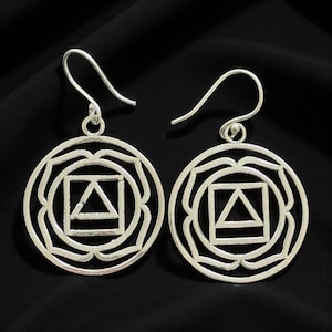 Op de afbeelding: Een paar zilverkleurige oorbellen. Elke oorbel heeft een cirkelvormig ontwerp met een geometrische vierkante en driehoekige vorm in het midden, omgeven door een bloemachtig patroon. De oorbellen hebben een haaksluiting.