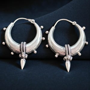 Peut inclure: Une paire de boucles d'oreilles créoles argentées. Chaque boucle d'oreille présente un design en forme de croissant avec de petits ornements ronds sur le bord extérieur. Un élément décoratif est suspendu au centre, avec une bande texturée et un pendentif pointu en forme de goutte.