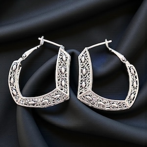 Peut inclure: Une paire de boucles d'oreilles créoles carrées argentées. Les boucles d'oreilles présentent un motif ajouré avec des motifs floraux et de volutes complexes. Les boucles d'oreilles sont présentées sur une surface noire.
