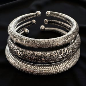 Op de afbeelding: Vier zilverkleurige armbanden met gedetailleerde patronen. De bovenste armband heeft een bloemenpatroon, de middelste een gedetailleerd bloemendesign en de onderste een gestructureerd, gestippeld patroon. De armbanden zijn gestapeld op een zwarte achtergrond.