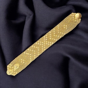 Pode incluir: Uma pulseira de tom dourado com uma banda retangular larga de padrão de malha densa e texturizada. A pulseira inclui dois fechos metálicos circulares estampados numa extremidade e um fecho de botão dourado, liso e arredondado, na outra.