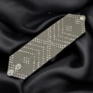 Peut inclure: Bracelet manchette argenté avec un motif tissé texturé. Le bracelet est de forme rectangulaire avec des bords arrondis et deux fermetures à boutons décoratifs. Le bracelet est présenté sur un fond de satin noir.