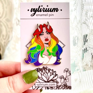 Könnte beinhalten: Eine Regenbogen-Emaille-Anstecknadel mit einer Comic-Frau mit Hörnern und einer regenbogenfarbenen Frisur. Die Anstecknadel befindet sich auf einer weißen Karte mit dem Text "vylirium. enamel pin" und "vylirium.com".