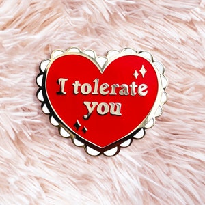 Puede incluir: Pin esmaltado en forma de corazón con centro rojo y borde dorado. El pin presenta el texto "I tolerate you" en letras doradas, junto con dos pequeñas chispas en forma de diamante. El pin tiene un borde festoneado.