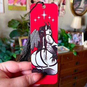 May include: Red bookmark with a black and white illustration of a demon with wings and a snake. The demon has a 666 tattoo on their leg and is wearing a black choker. The bookmark has a black tassel.