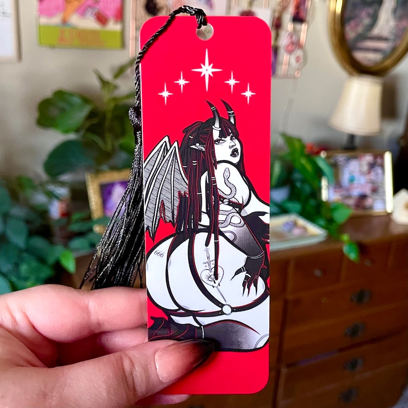 May include: Red bookmark with a black and white illustration of a demon with wings and a snake. The demon has a 666 tattoo on their leg and is wearing a black choker. The bookmark has a black tassel.