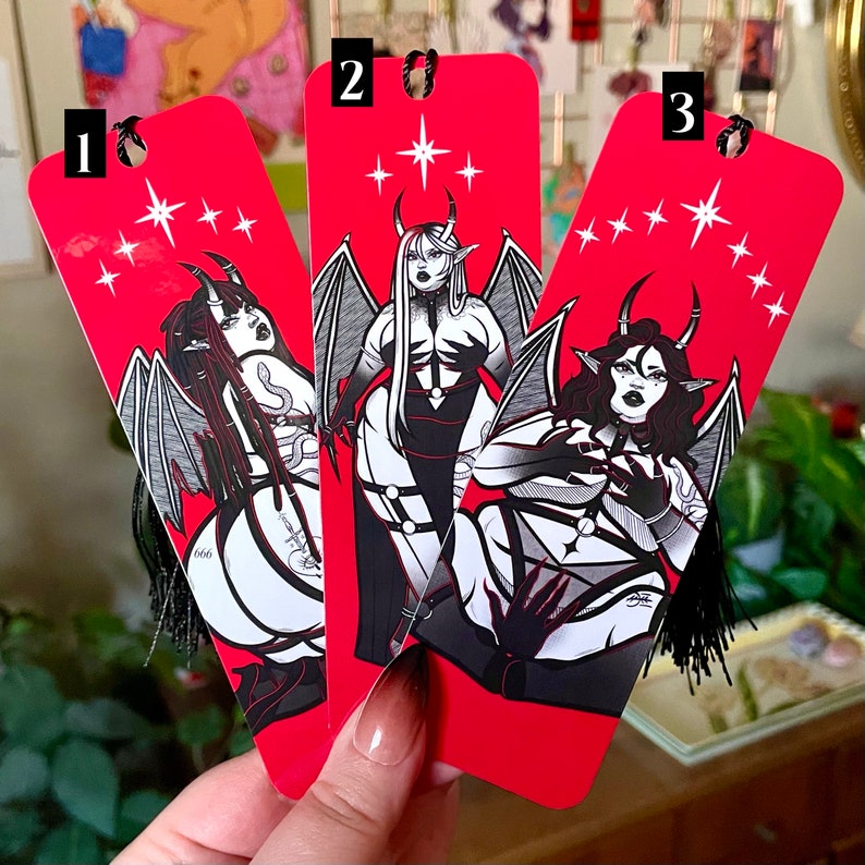 May include: Three red bookmarks with black and white illustrations of women with horns and wings. The bookmarks have a red background with white stars and a black tassel.