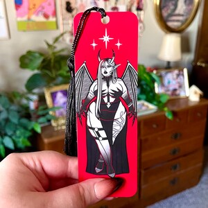 May include: Red bookmark with a black and white illustration of a woman with wings and horns. The woman is wearing a long black dress and has a snake wrapped around her leg. The bookmark has a black tassel.