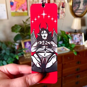 May include: A red bookmark with a black and white illustration of a demon with horns and wings. The demon is wearing a harness and has a halo above its head. The background is red with white stars.