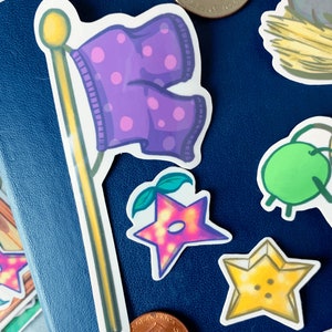May include: A set of five stickers featuring a purple flag with polka dots, a pink star with a green stem, a yellow star with a button, and two green apples. The stickers are on a blue background.