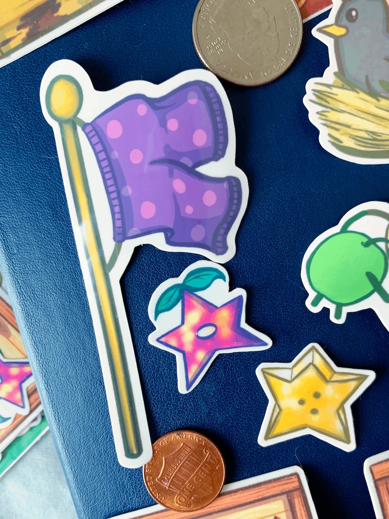 May include: A set of five stickers featuring a purple flag with polka dots, a pink star with a green stem, a yellow star with a button, and two green apples. The stickers are on a blue background.