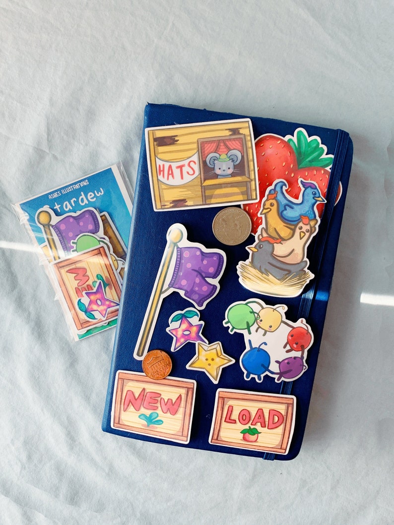 May include: A blue notebook with various stickers on it. The stickers include a strawberry, a bird, a mouse, a hat shop, a star, a flag, a coin, and a sign that says "NEW" and "LOAD".