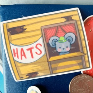 May include: A yellow and brown wooden shop with a red curtain and a sign that says "HATS". A grey mouse with a green hat is looking out of the shop window.