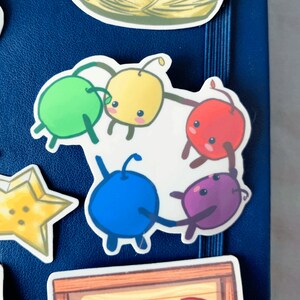 May include: Five colourful cartoon characters holding hands in a circle. The characters are green, yellow, red, blue, and purple. The characters are on a white background.