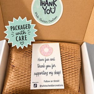 Puede incluir: Una caja de cart&oacute;n marr&oacute;n con un paquete envuelto. El paquete est&aacute; sellado con una pegatina que dice "Thank You" y "Packaged with Care". Una tarjeta con un gr&aacute;fico de donut dice "&iexcl;Divi&eacute;rtete y gracias por apoyar mi tienda!" e incluye un handle de TikTok.