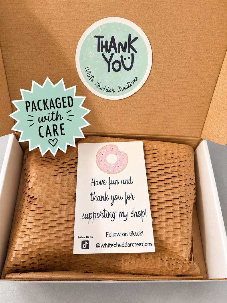 Puede incluir: Una caja de cart&oacute;n marr&oacute;n con un paquete envuelto. El paquete est&aacute; sellado con una pegatina que dice "Thank You" y "Packaged with Care". Una tarjeta con un gr&aacute;fico de donut dice "&iexcl;Divi&eacute;rtete y gracias por apoyar mi tienda!" e incluye un handle de TikTok.