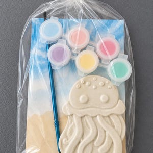 Puede incluir: Una bolsa de pl&aacute;stico transparente contiene un kit para pintar tu propia medusa. El kit incluye una pieza de cer&aacute;mica beige con forma de medusa, seis botes de pintura peque&ntilde;os en colores pastel y un pincel azul. El fondo presenta una escena de playa.
