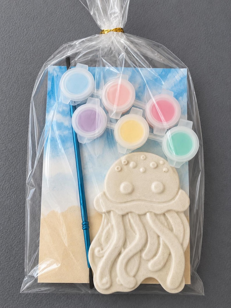 Puede incluir: Una bolsa de pl&aacute;stico transparente contiene un kit para pintar tu propia medusa. El kit incluye una pieza de cer&aacute;mica beige con forma de medusa, seis botes de pintura peque&ntilde;os en colores pastel y un pincel azul. El fondo presenta una escena de playa.