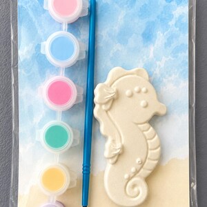 Puede incluir: Un kit de pintura de caballito de mar. El kit incluye un caballito de mar de cer&aacute;mica, un pincel azul y seis botes de pintura en rosa, azul, rosa, verde, amarillo y morado. El fondo presenta un dise&ntilde;o de acuarela de playa y cielo.