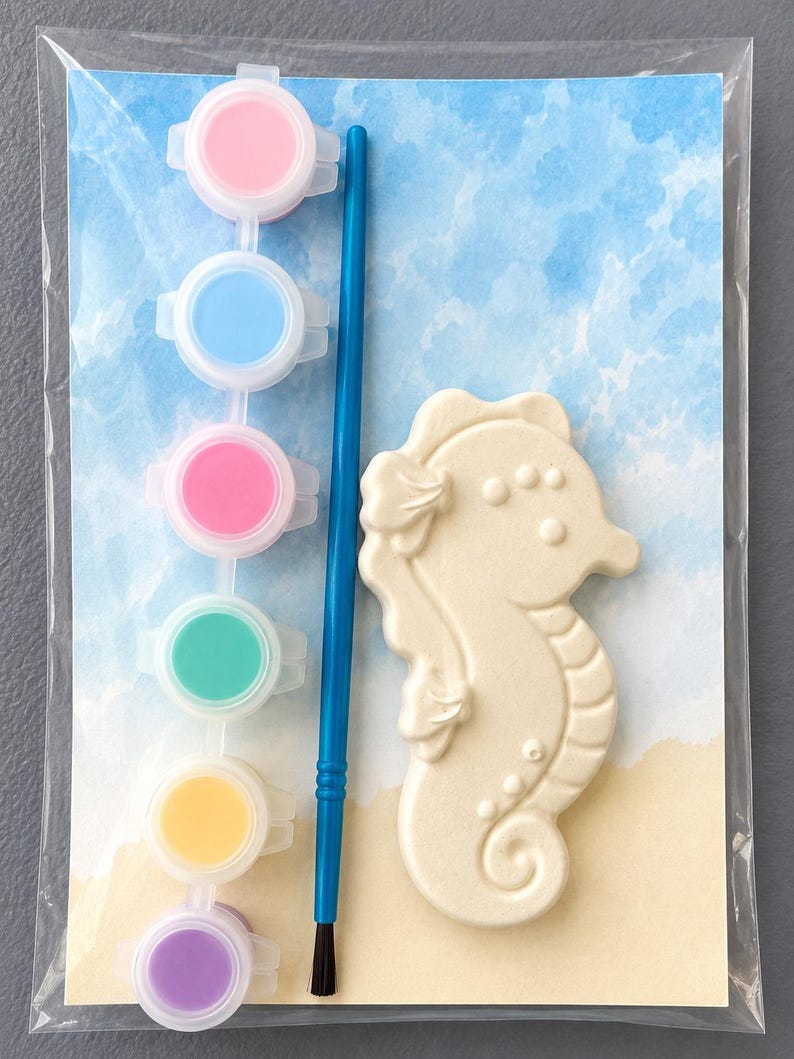Puede incluir: Un kit de pintura de caballito de mar. El kit incluye un caballito de mar de cer&aacute;mica, un pincel azul y seis botes de pintura en rosa, azul, rosa, verde, amarillo y morado. El fondo presenta un dise&ntilde;o de acuarela de playa y cielo.