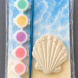Puede incluir: Un kit de pintura por n&uacute;meros con un dise&ntilde;o de concha. El kit incluye un lienzo en forma de concha, un pincel azul y seis botes de pintura en varios colores: verde, amarillo, morado, rosa, rosa claro y azul. El fondo representa una escena de playa.