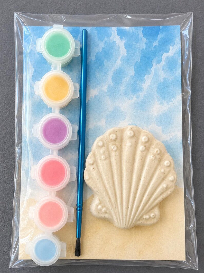 Puede incluir: Un kit de pintura por n&uacute;meros con un dise&ntilde;o de concha. El kit incluye un lienzo en forma de concha, un pincel azul y seis botes de pintura en varios colores: verde, amarillo, morado, rosa, rosa claro y azul. El fondo representa una escena de playa.