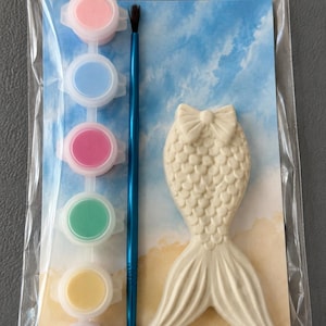 Puede incluir: Kit de manualidades de yeso con forma de cola de sirena con un pincel y botes de pintura en rosa, azul, rojo, verde, amarillo y morado. El kit est&aacute; empaquetado en una bolsa de pl&aacute;stico transparente, con un fondo azul y beige.