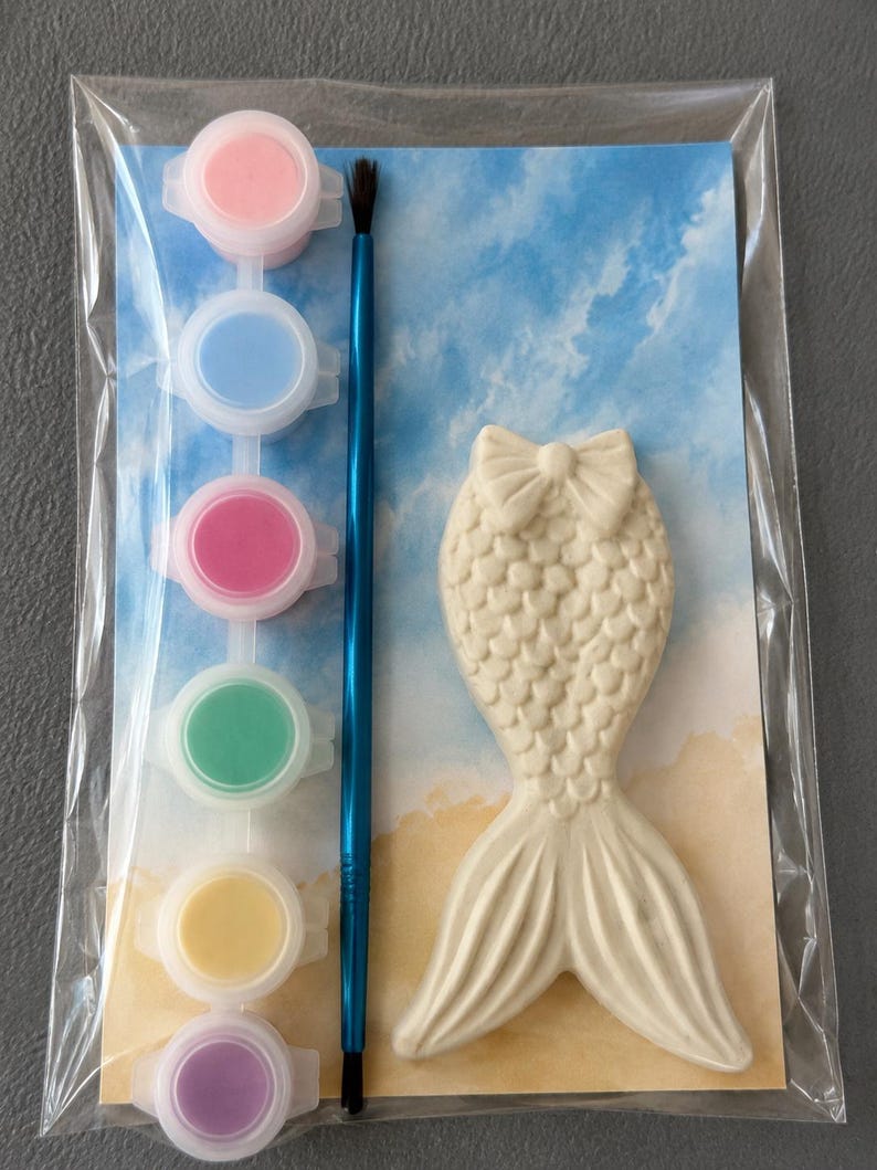 Puede incluir: Kit de manualidades de yeso con forma de cola de sirena con un pincel y botes de pintura en rosa, azul, rojo, verde, amarillo y morado. El kit est&aacute; empaquetado en una bolsa de pl&aacute;stico transparente, con un fondo azul y beige.