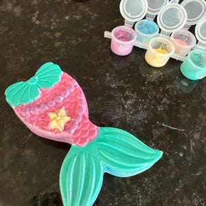 Puede incluir: Un proyecto de manualidades con forma de cola de sirena, pintado en tonos rosa, verde y amarillo. La cola presenta un lazo, escamas y una estrella. En el fondo se ven peque&ntilde;os botes de pintura de varios colores, lo que sugiere un kit de manualidades.