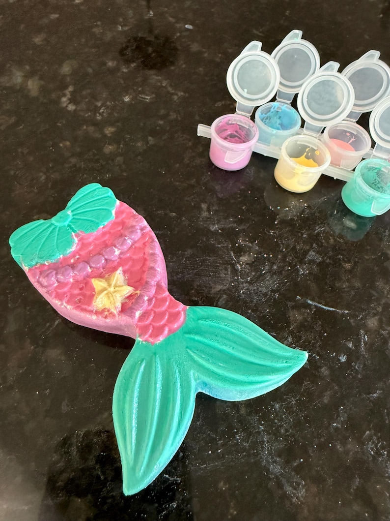 Puede incluir: Un proyecto de manualidades con forma de cola de sirena, pintado en tonos rosa, verde y amarillo. La cola presenta un lazo, escamas y una estrella. En el fondo se ven peque&ntilde;os botes de pintura de varios colores, lo que sugiere un kit de manualidades.