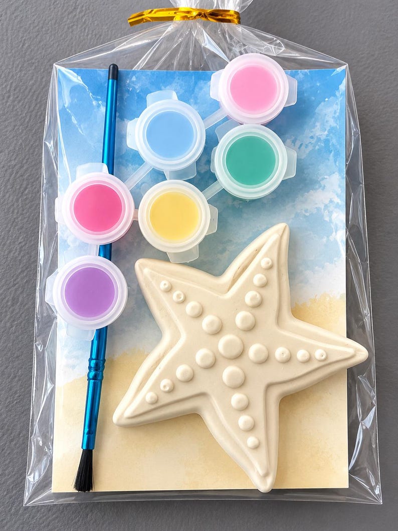 Puede incluir: Un kit de pintura de estrella de mar en una bolsa transparente. El kit incluye una estrella de mar crema, un pincel azul y seis botes de pintura rosa, azul, verde, amarillo, rosa y morado. El fondo muestra una escena de playa.