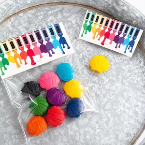 May include: A clear plastic bag containing eleven seashell-shaped crayons in various colors including red, orange, yellow, green, blue, purple, and black. Two rectangular paper toppers feature a colorful graphic print of dripping paint tubes.