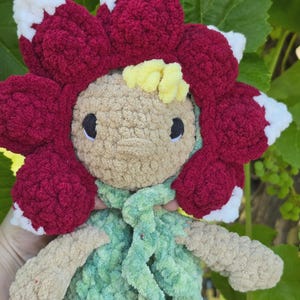 May include: A plush doll crafted from soft yarn, featuring a tan face with black eyes and yellow hair. The doll wears a large, textured hood shaped like a deep red flower with white-tipped petals and a light green textured vest with a scarf.