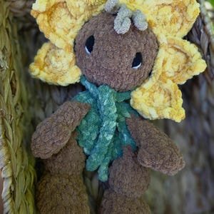 May include: A plush toy shaped like a sunflower with a brown head, yellow petals, and a green textured collar. The toy features a brown body with matching arms and legs, and small black eyes with white accents, crafted from soft, fuzzy yarn material.