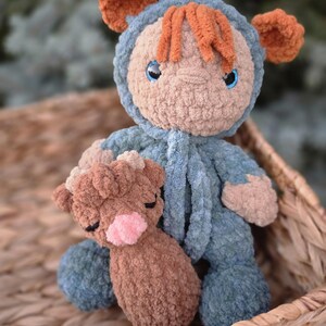 May include: A handmade crochet plush doll wearing a blue hooded onesie with bear ears and orange hair, featuring blue eyes. The doll is holding a smaller, brown crochet plush animal with a pink nose and closed eyes, crafted from soft yarn.