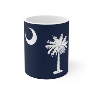 May include: White ceramic mug displaying the South Carolina state flag. The design features a white crescent moon and a white palmetto tree on a navy blue background. The mug has a white interior.