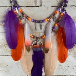 May include: A handcrafted necklace featuring layers of orange, purple, and silver beads. The necklace is adorned with colorful feathers in shades of purple, orange, and cream, and a pendant with a desert landscape design. The necklace is a bohemian style.