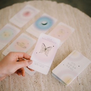 May include: A set of affirmation cards with pastel colors and a butterfly design. The card in the foreground reads "New Beginnings" with the text "Change is coming | Rebirth | I am you". The card is being held by a hand. The other cards are blurred in the background. A small book with the text "It's Already Yours" is also visible.