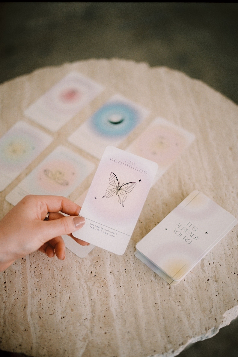 May include: A set of affirmation cards with pastel colors and a butterfly design. The card in the foreground reads "New Beginnings" with the text "Change is coming | Rebirth | I am you". The card is being held by a hand. The other cards are blurred in the background. A small book with the text "It's Already Yours" is also visible.