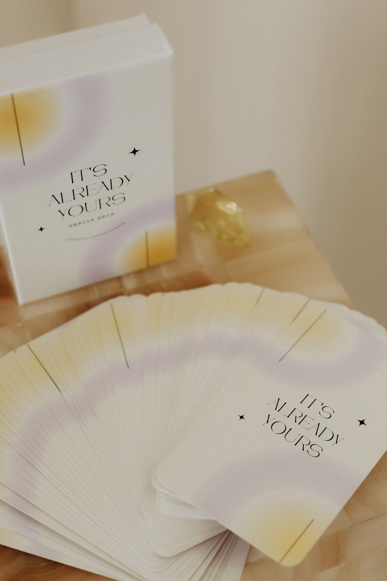May include: A deck of oracle cards with a soft, pastel color scheme. The cards are fanned out and the top card reads "It's Already Yours" in black text. The cards are on a light brown wooden surface.