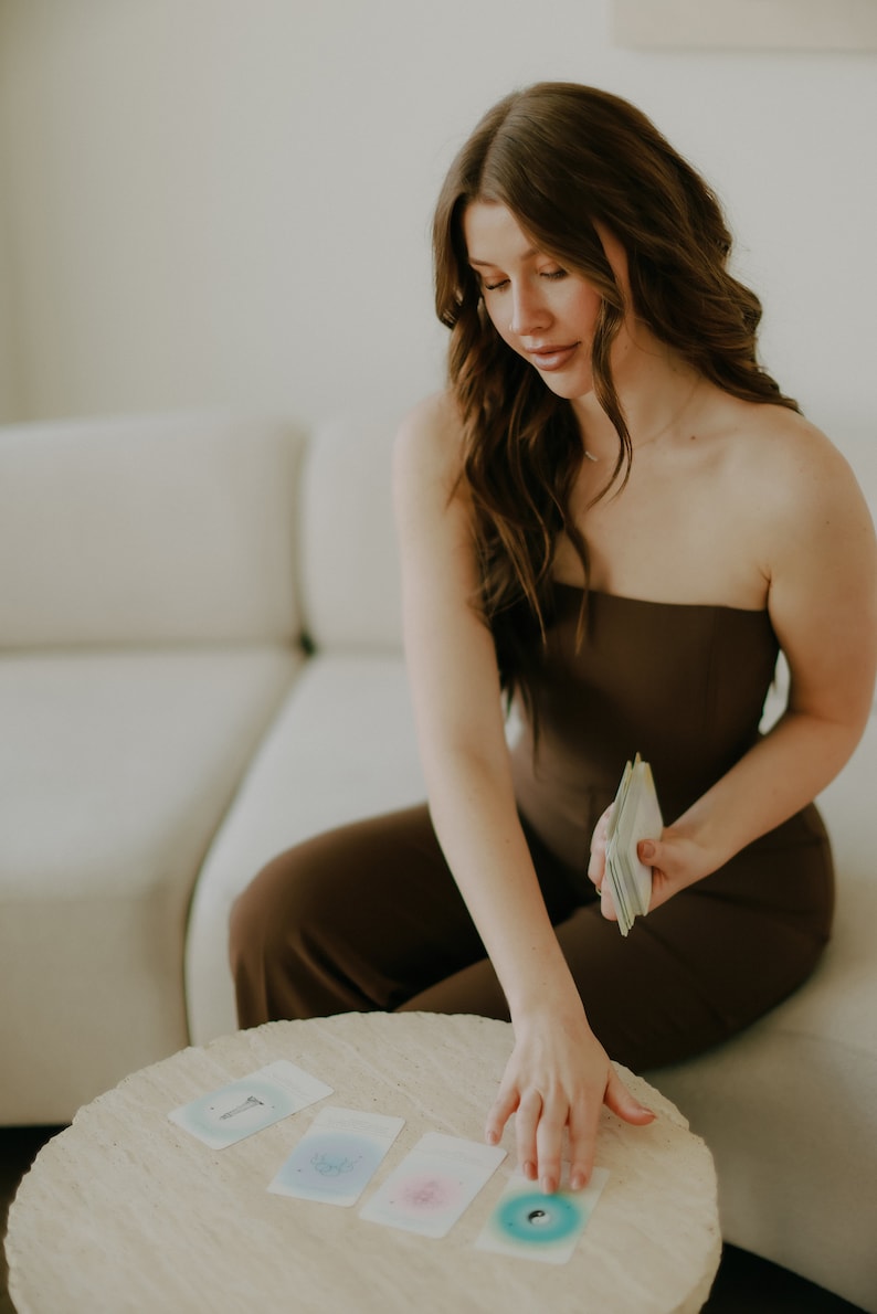 May include: A woman in a brown strapless jumpsuit sits on a white couch and lays out cards on a round, light brown table. The cards have colorful designs and are spread out in a row.