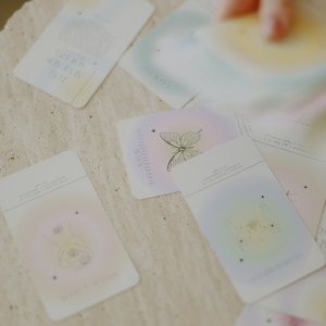 May include: A set of pastel-colored oracle cards with illustrations of butterflies, stars, and other celestial imagery. The cards have text on them, including the words "Realignments" and "Reconnecting".